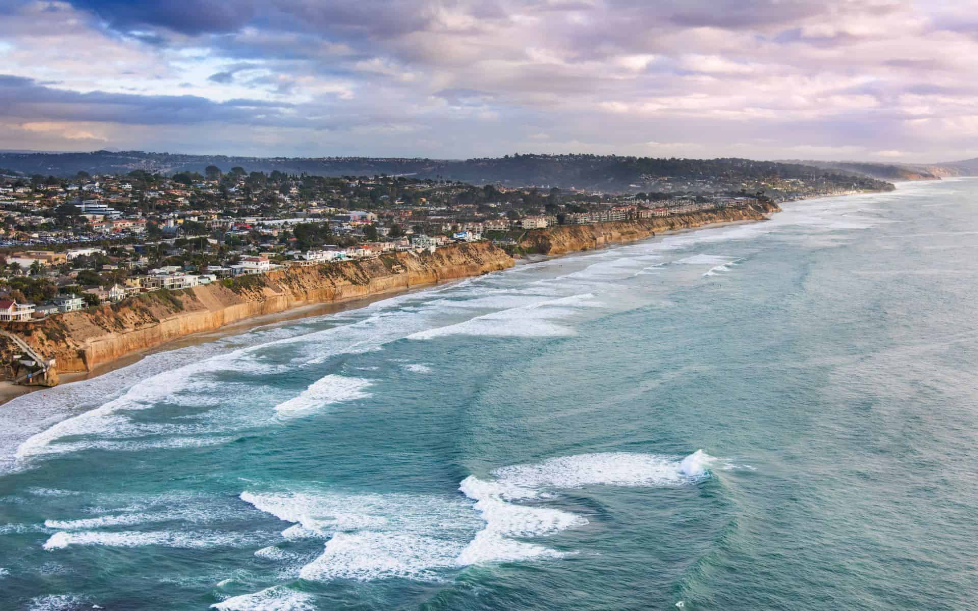 Aerial view of the Solana Beach coastline with waves breaking along the shore, golden cliffs lined with homes and trees, and a pastel sky above.