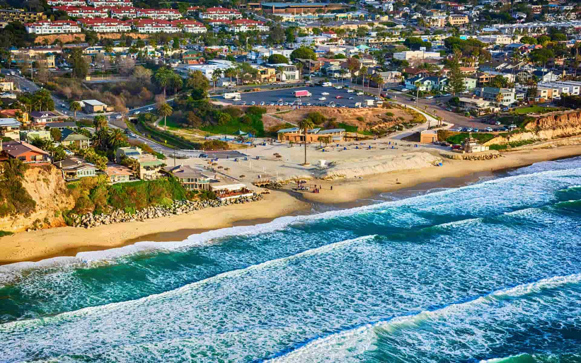 Aerial view of Encinitas beach in San Diego County, California, featuring sandy shore with gentle waves, cliffs, green vegetation, residential neighborhoods with red roofs, parking lot, pathways, and people on the beach. ​