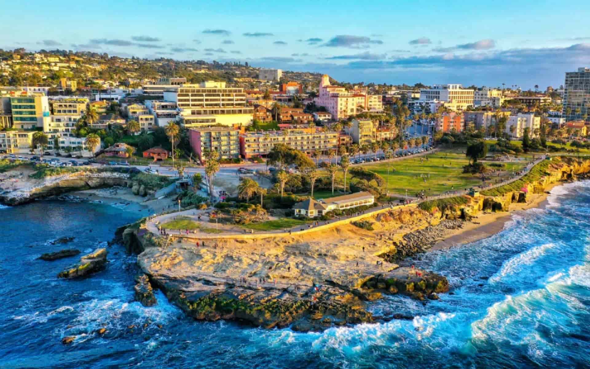Aerial drone view of La Jolla Cove in San Diego, featuring crashing waves on rocky cliffs, sandy beach, colorful buildings, high-rise tower, and hillside neighborhoods under partly cloudy skies.
