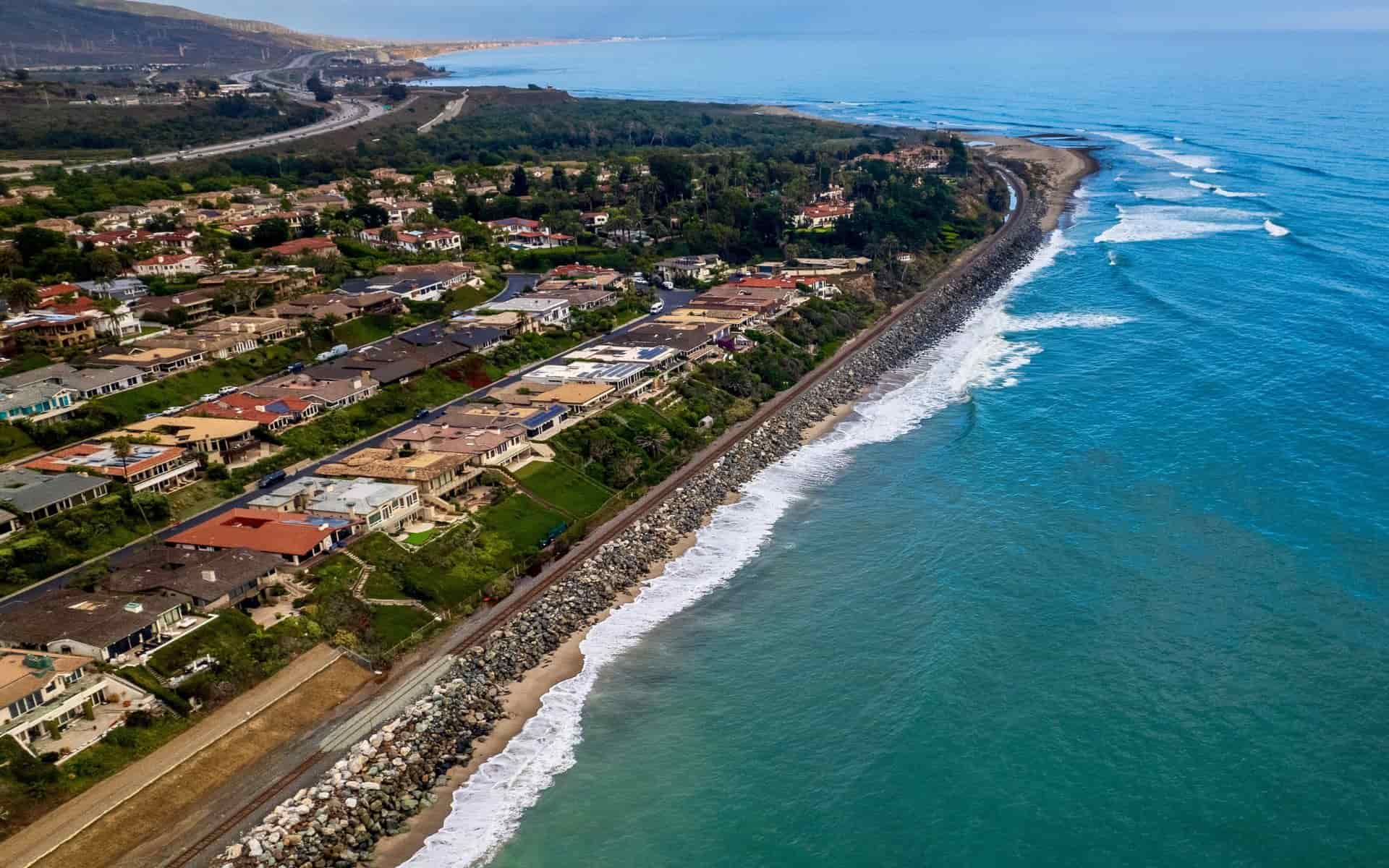 Aerial view of San Clemente coastal area, showing residential neighborhoods with red-roofed homes along the rocky shoreline, Pacific Ocean waves, highway, and green hills under clear skies.