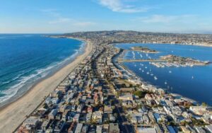 Aerial view of Mission Beach in San Diego showing a narrow peninsula between the Pacific Ocean and Mission Bay, lined with beachfront homes, marinas, and boats on a clear day.