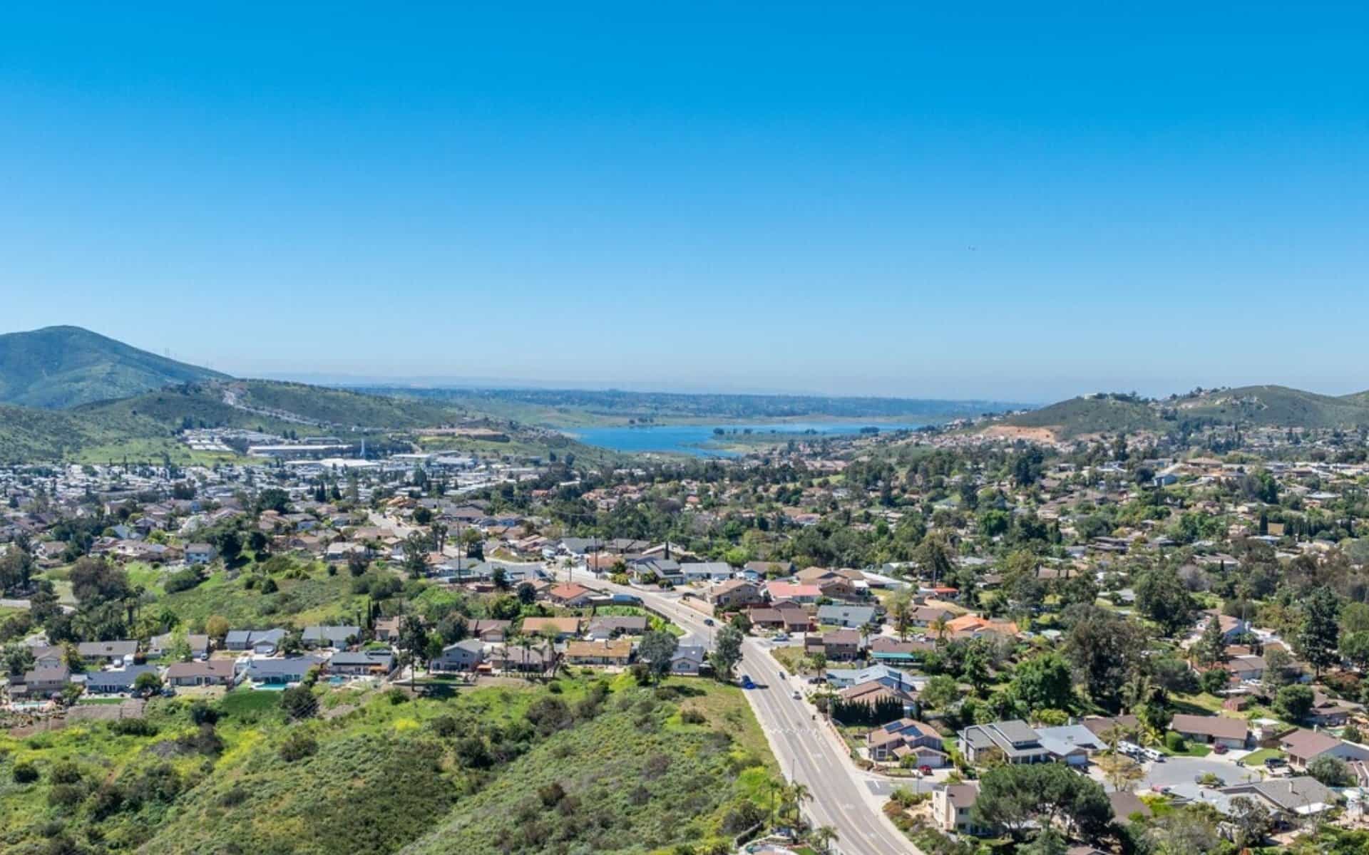 Aerial view of a suburban neighborhood in Spring Valley, California, with houses and streets in the foreground, green hills and a small lake in the mid-distance under a clear blue sky.