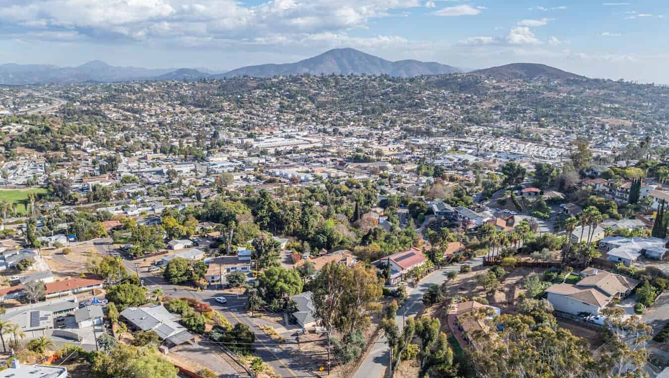 Wide aerial panorama of Spring Valley, California: dense residential neighborhoods in the foreground stretching to green hills and distant mountains beneath a partly cloudy sky