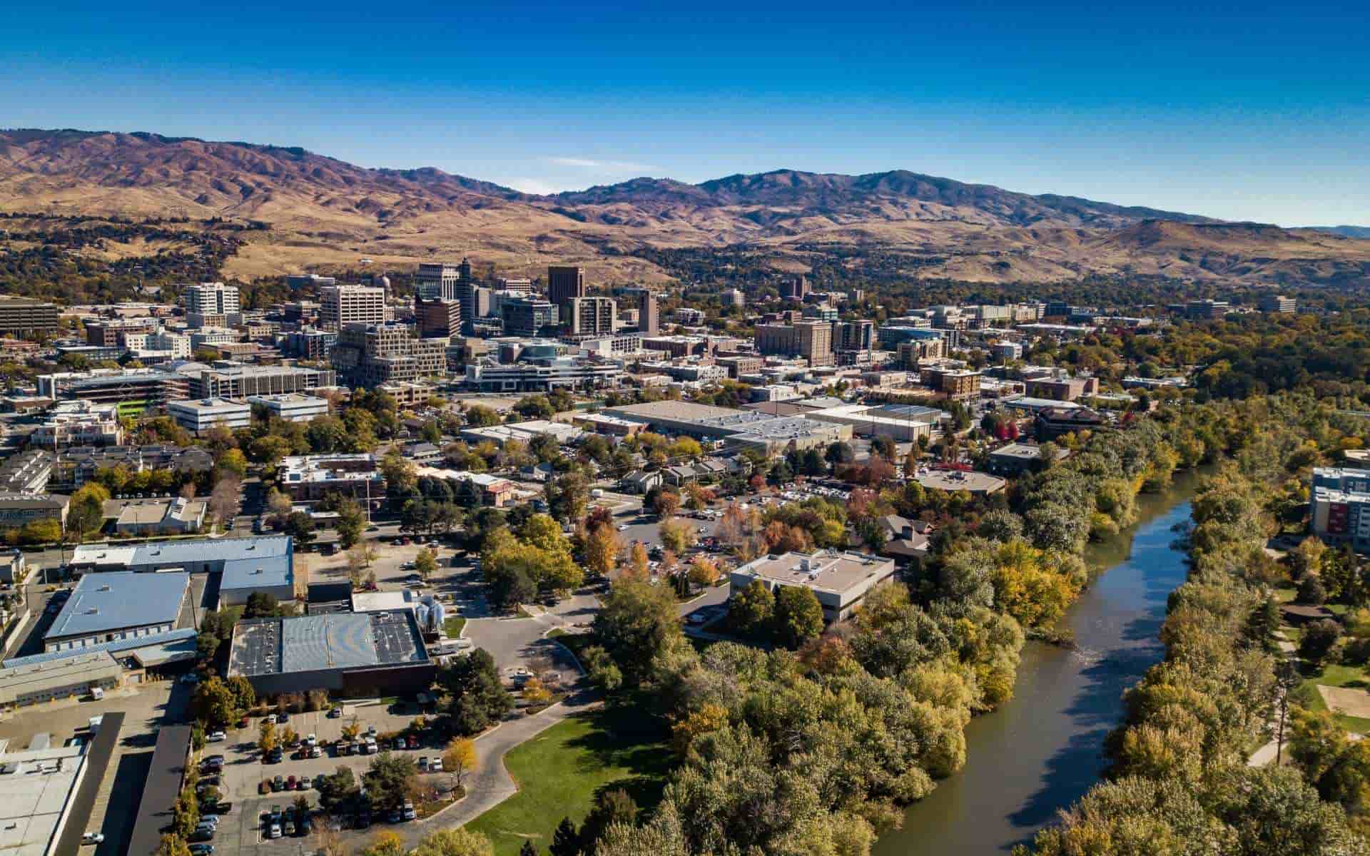 Aerial view of downtown Boise, the tree-lined Boise River, and surrounding foothills beneath a blue sky.