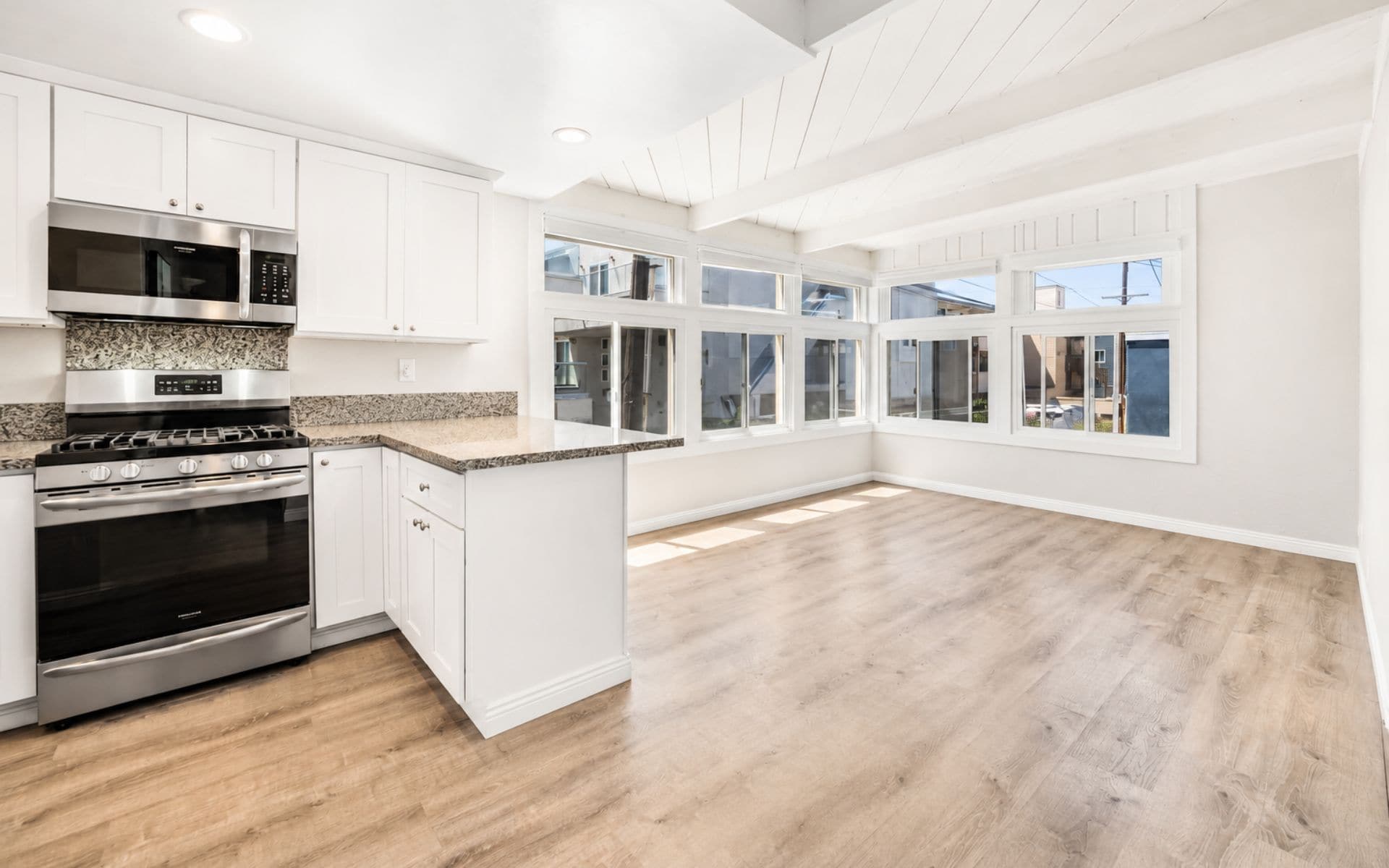 Unfurnished living area with wood flooring, large windows, and an open kitchen with white cabinets and stainless steel appliances.