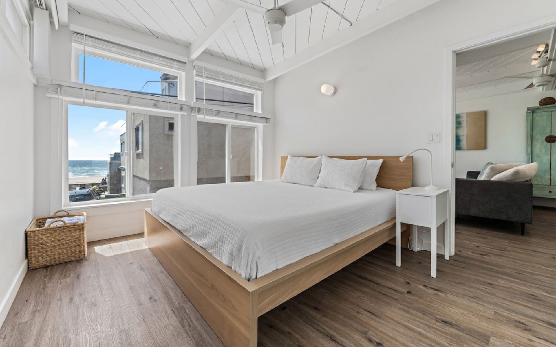 Bright bedroom with white bedding, a wooden bed frame, vaulted ceiling, and an ocean glimpse through large windows.