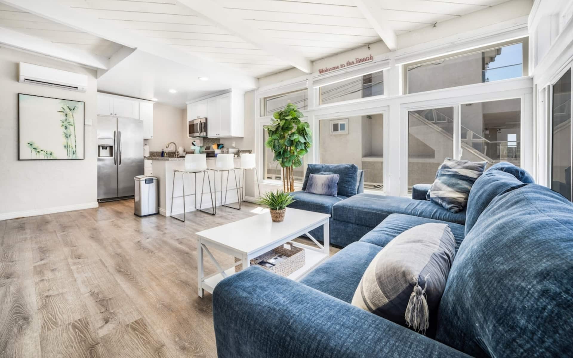 Bright furnished living room with a blue sectional sofa, white coffee table, large windows, and an open kitchen.